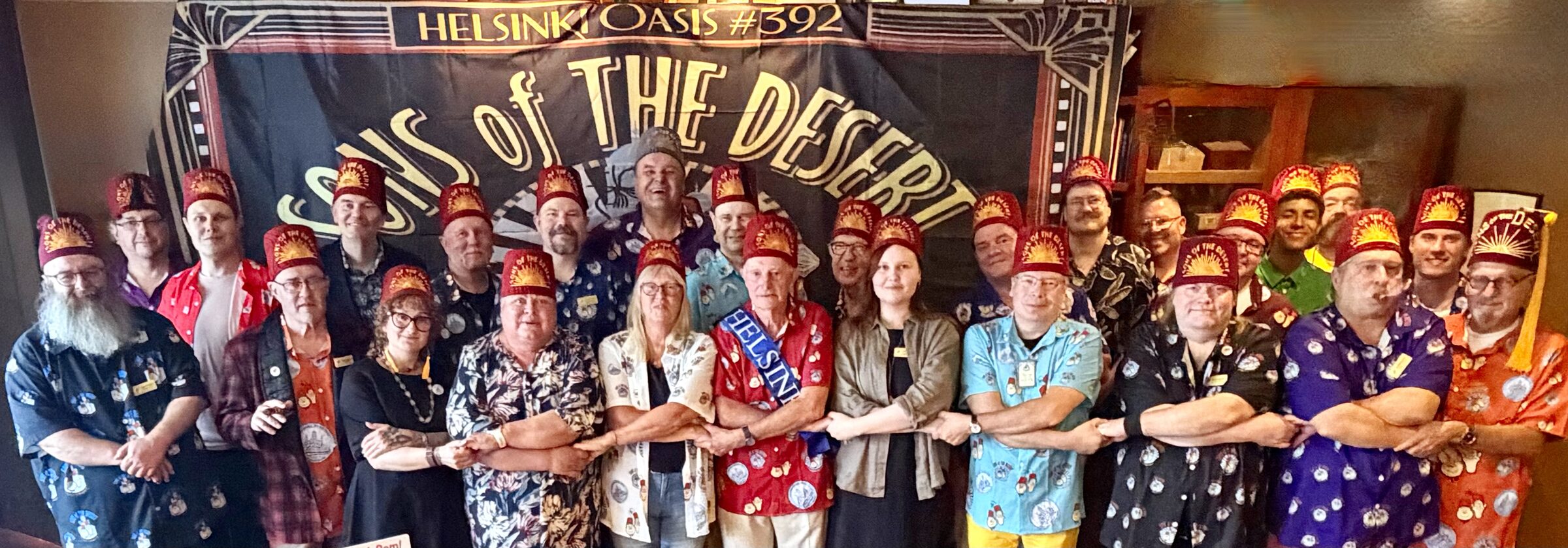 Helsinki Oasis #392:n jäsenet fetseissään ja Laurel ja Hardy -paidoissaan käsi kädessä Sons of the Desert -banderollin edessä Roger Robinsonin kanssa